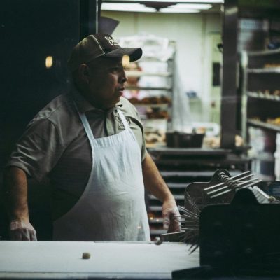 An adult male baker in an apron works in a donut shop kitchen, surrounded by baking equipment.