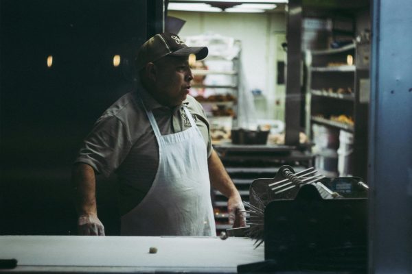 An adult male baker in an apron works in a donut shop kitchen, surrounded by baking equipment.