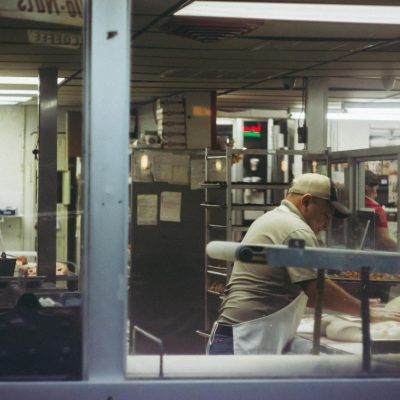 Baker working with dough in a commercial bakery kitchen, viewed through a glass window.