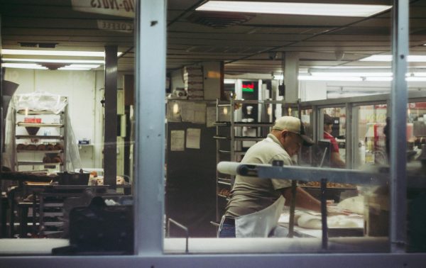 Baker working with dough in a commercial bakery kitchen, viewed through a glass window.