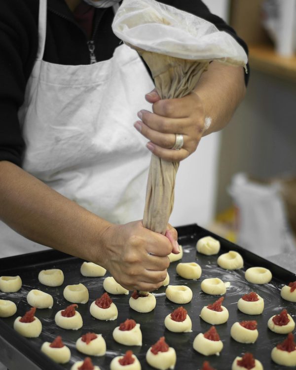 Close-up of hands piping jam into cookie dough in a bakery setup, showcasing pastry preparation.